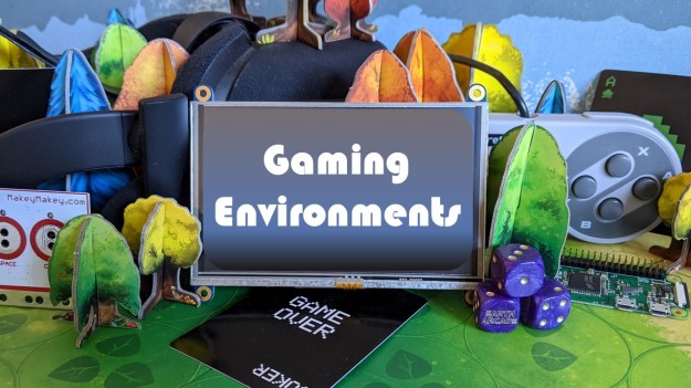 A screen with Gaming Environments written on it. It sits surrounded by gaming items, the green gaming board from Photosynthesis and green, yellow, orange, and blue trees. There is a gaming controller, VR headset and a Makey-makey.