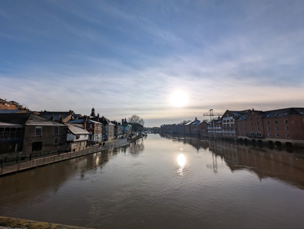 A view of the river Ouse taken from a bridge half way over its span. Buildings stand close to either side of the river. The sun is low in the blue sky, with whispy clouds, and reflects in the flat water surface.