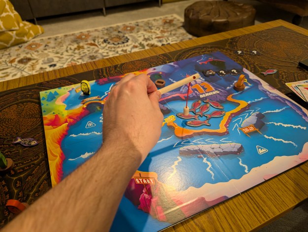 The 13 Beavers game board. Colourful cartoon style, with board squares along a rapid flowing river. A player's hand is using a small fishing rod to catch magnetic fish.