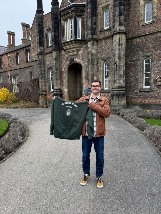 A photo of me outside the an old building holding up a York St John University hoodie.