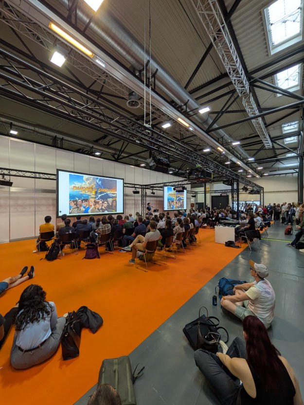 A few from the back of a presentation room. People sit on rows of chairs in front of large screens.